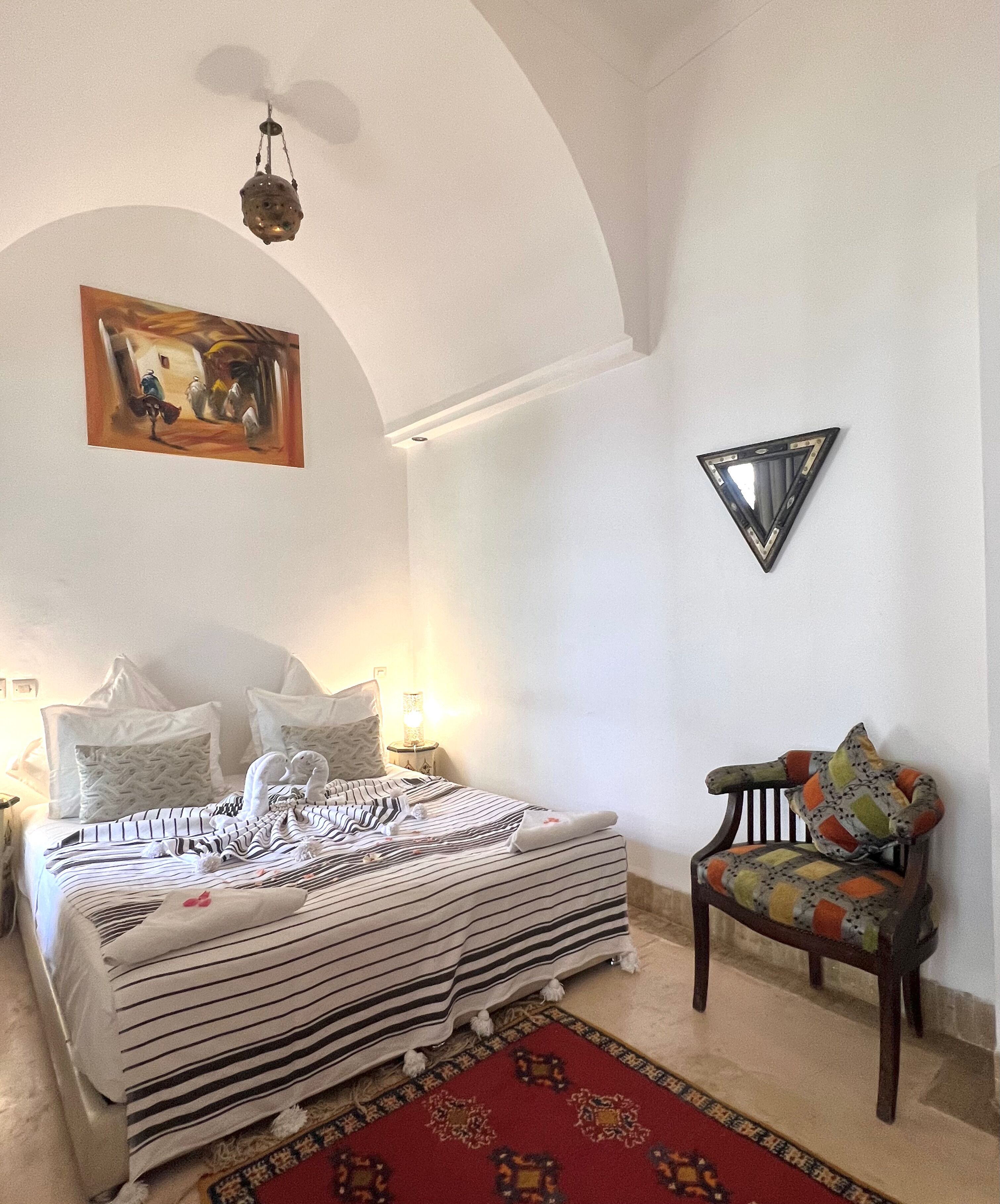 A bedroom with a bed, a chair, a painting, and a triangular mirror on the wall.