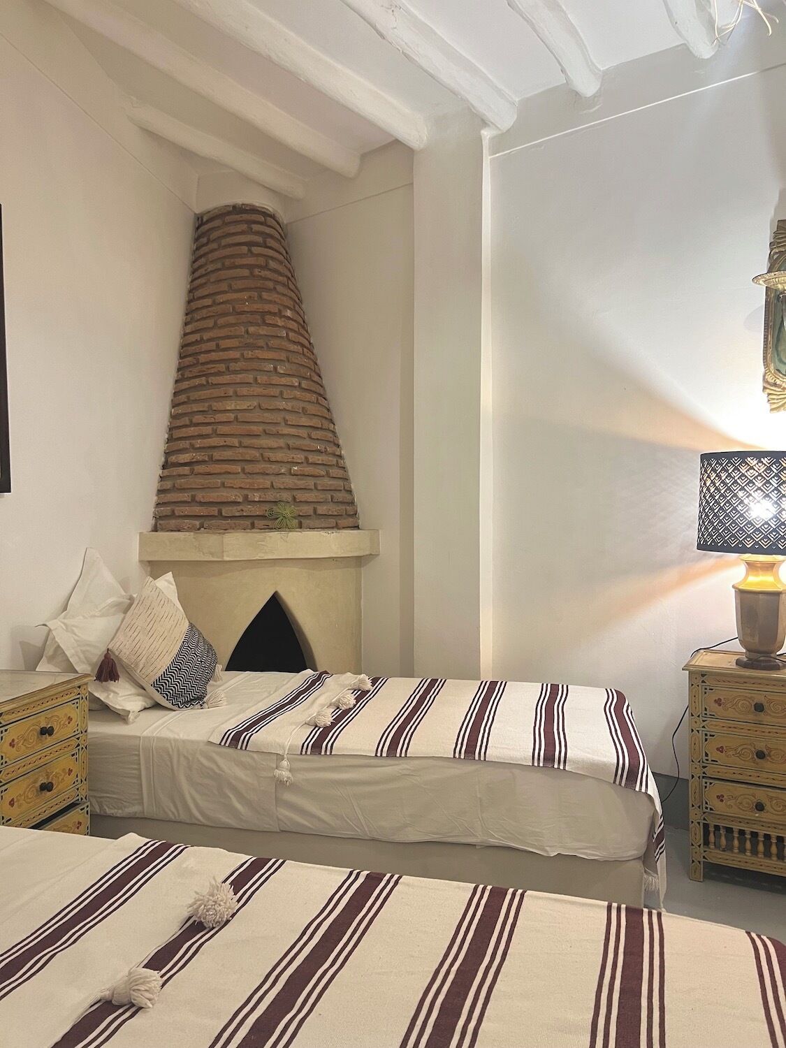 Un dormitorio con chimenea de ladrillo, una cama con ropa de cama a rayas, una mesita de noche con lámpara y un mueble auxiliar con cajones.