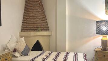 Un dormitorio con chimenea de ladrillo, una cama con ropa de cama a rayas, una mesita de noche con lámpara y un mueble auxiliar con cajones.