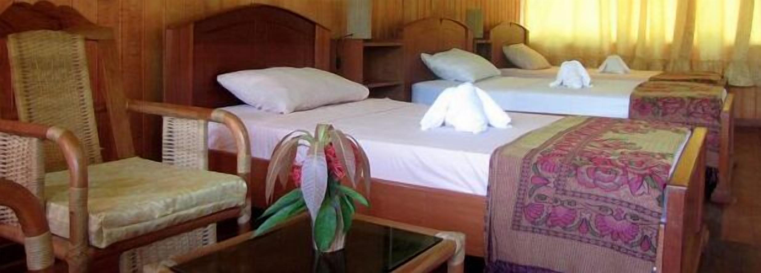Camera d'albergo con due letti, poggiateste in legno e una sedia con una pianta verde.