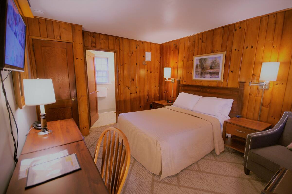 A hotel room with wooden paneling, a bed with white linens, two bedside tables with lamps, a desk with a lamp, a chair, and a television on the wall.