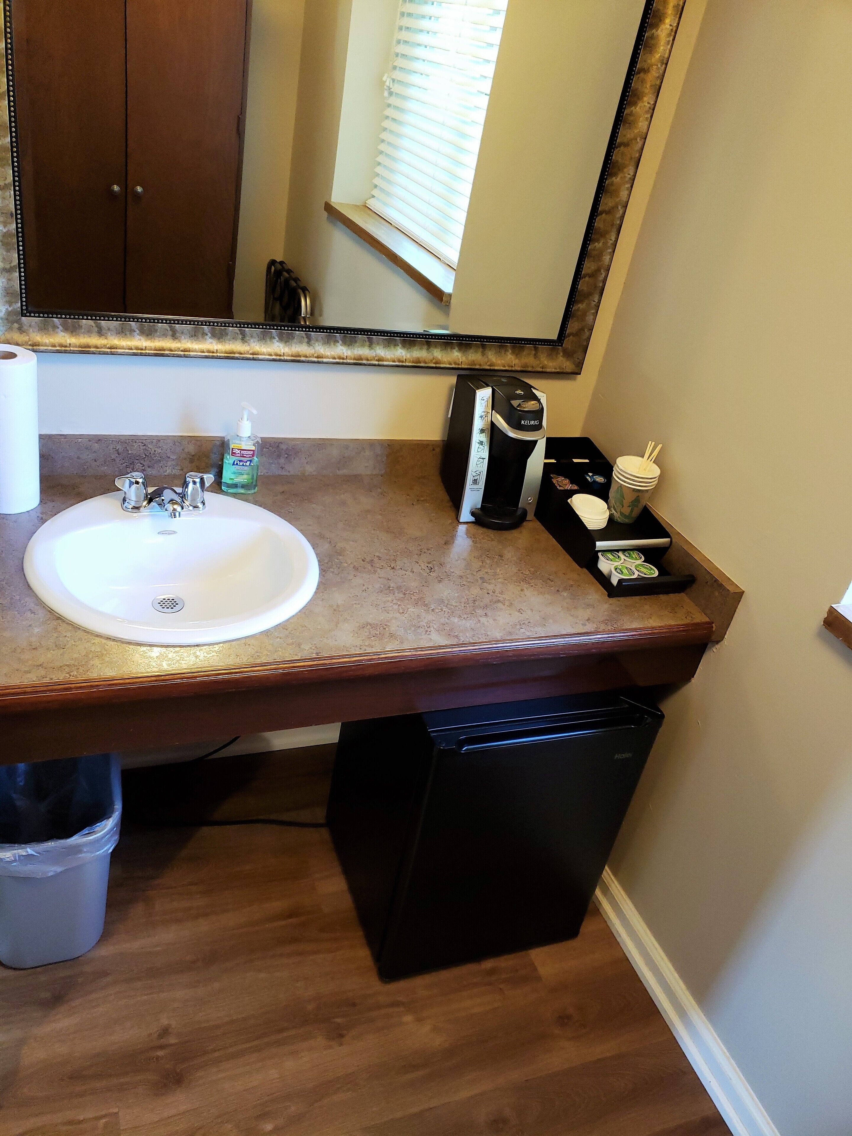A bathroom with a white sink, a mirror, a coffee maker, and a trash bin.