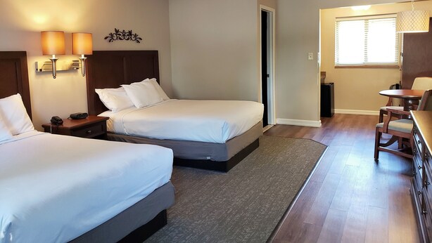 A hotel room with two beds, a desk, and a chair. There is a door leading to another room and a window with blinds.