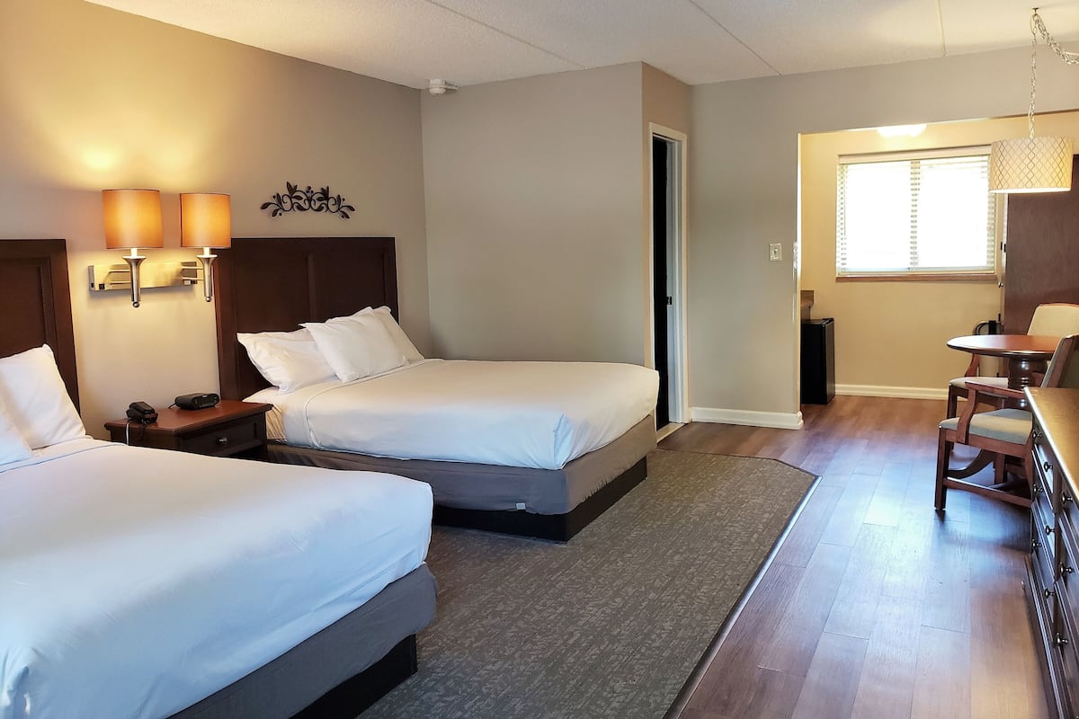 A hotel room with two beds, a desk, and a chair. There is a door leading to another room and a window with blinds.