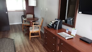 A hotel room with a wooden dining table and chairs, a coffee maker, a desk with drawers, and a large mirror.
