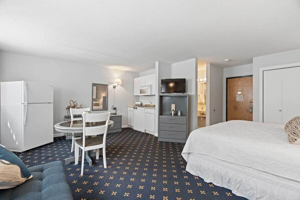 A compact hotel room with a bed, a small kitchenette, a dining area with a table and chairs, and a bathroom.