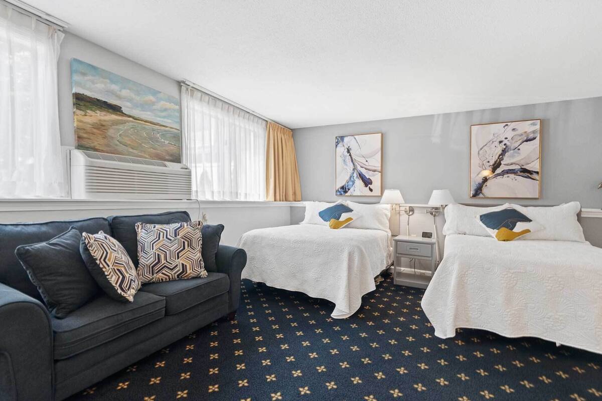 A hotel room with a dark blue carpet, a grey sofa with patterned cushions, two beds with white linens, and artwork on the walls.