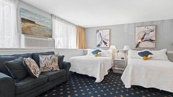 Une chambre d’hôtel avec un tapis bleu foncé, un canapé gris agrémenté de coussins à motifs, deux lits avec du linge de lit blanc et des œuvres d’art accrochées aux murs.