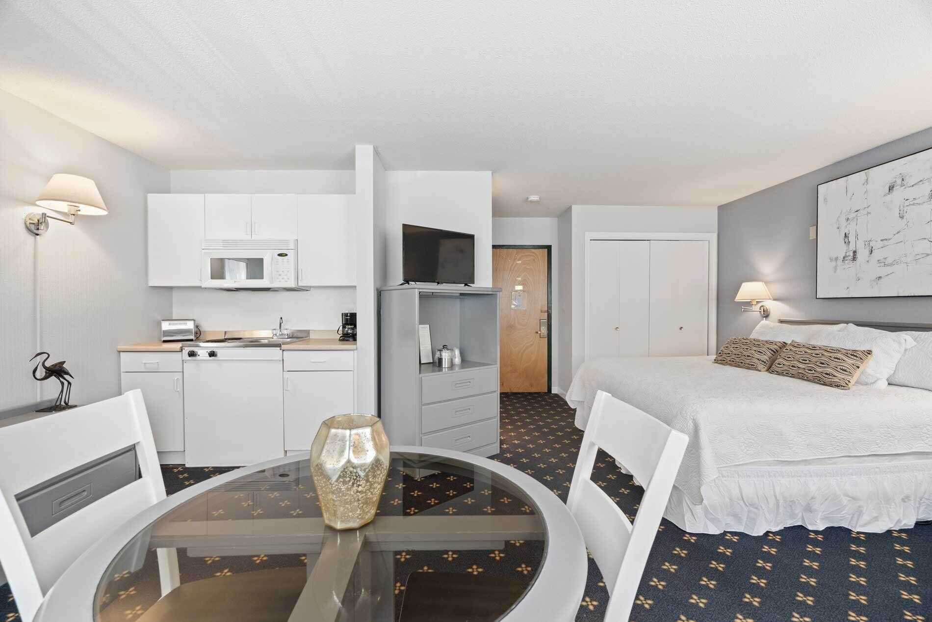 A hotel room with a bed, a desk with a chair, a small kitchenette, and a TV.