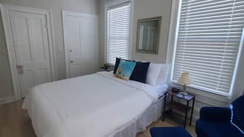 A bedroom with a large bed, two bedside tables, a chair, and a window with blinds.