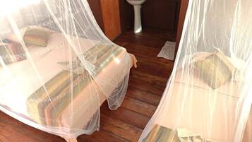 A room with two beds covered by sheer curtains, wooden flooring, and a white pedestal toilet.