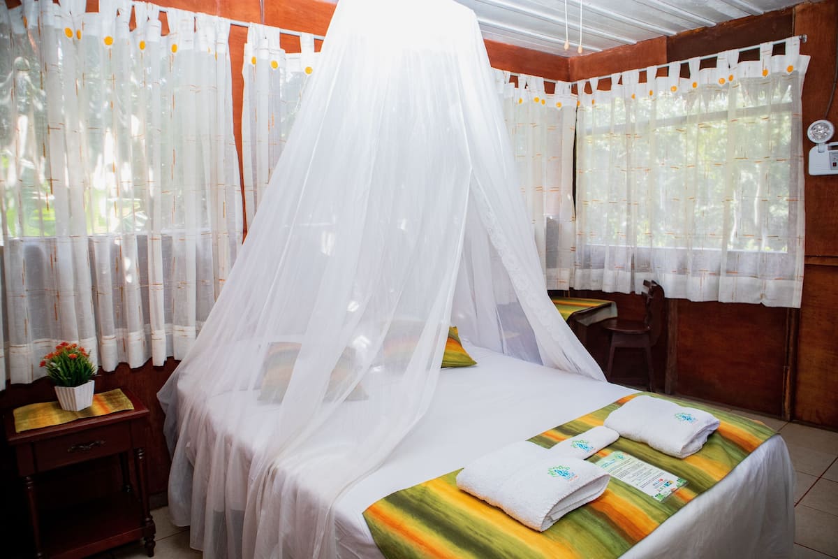 A room with a bed covered by a white canopy, a small table with a plant, and a chair.