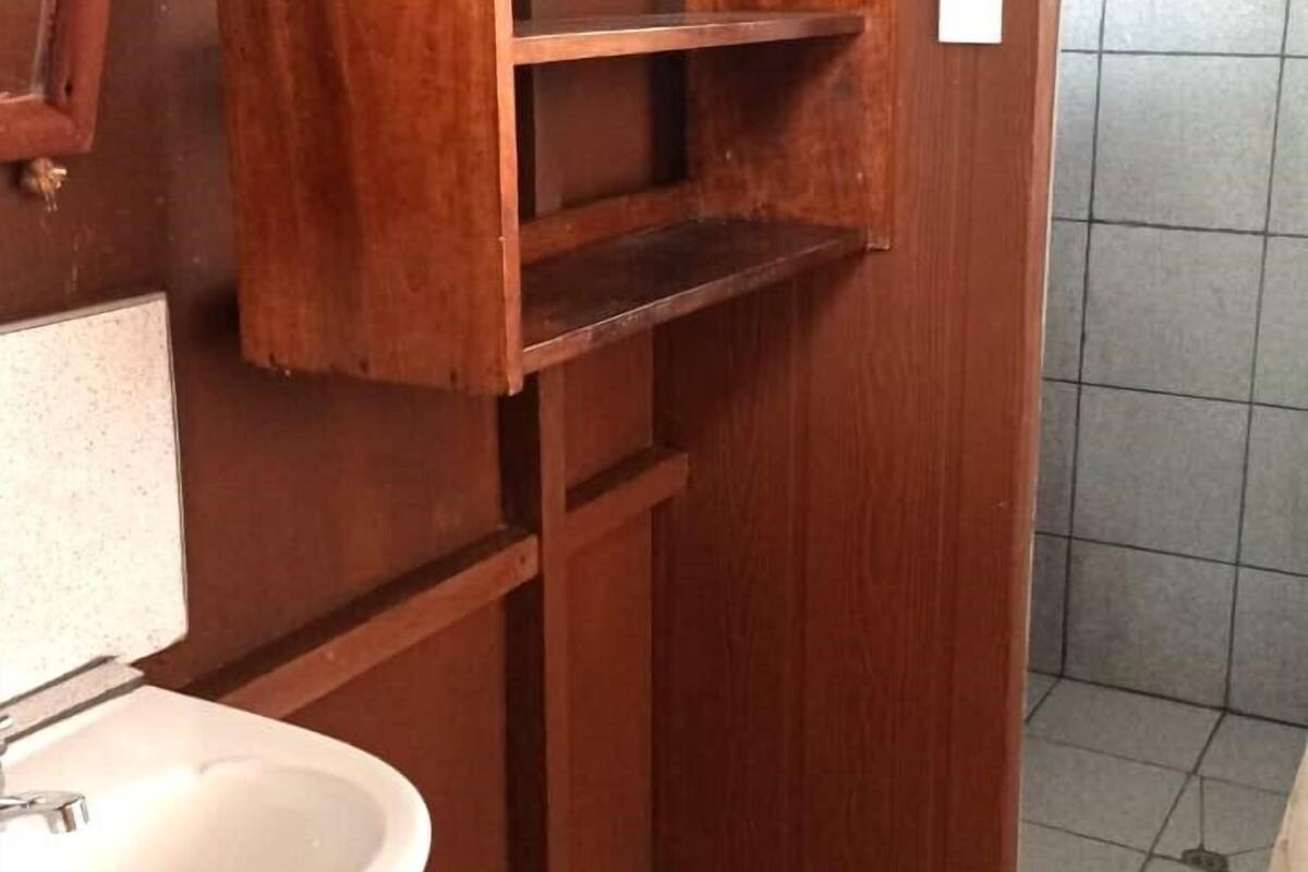 A wooden bathroom with a white sink, a wooden cabinet with open shelves, and a tiled shower area.