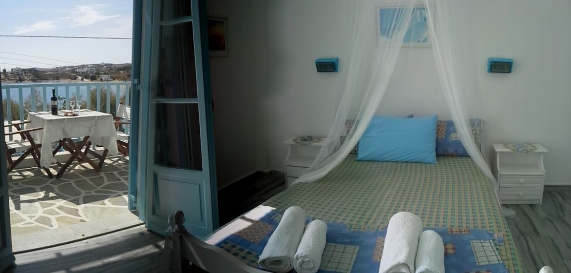 A bedroom with a bed under a canopy, a small bedside table, and a nightstand. There is a balcony with a table and chairs outside.