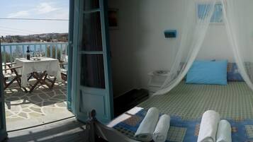 A bedroom with a bed under a canopy, a small bedside table, and a nightstand. There is a balcony with a table and chairs outside.