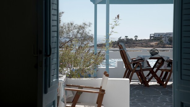 Vista desde una habitación con ventana a un patio. El patio tiene una mesa, sillas, una planta pequeña y vistas al mar y edificios al fondo.