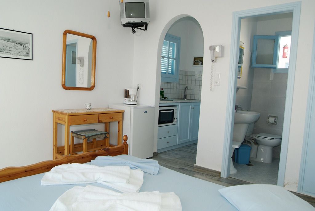 A bedroom with a bed, a wooden bedside table, a small desk, a mirror, and a television on the wall.