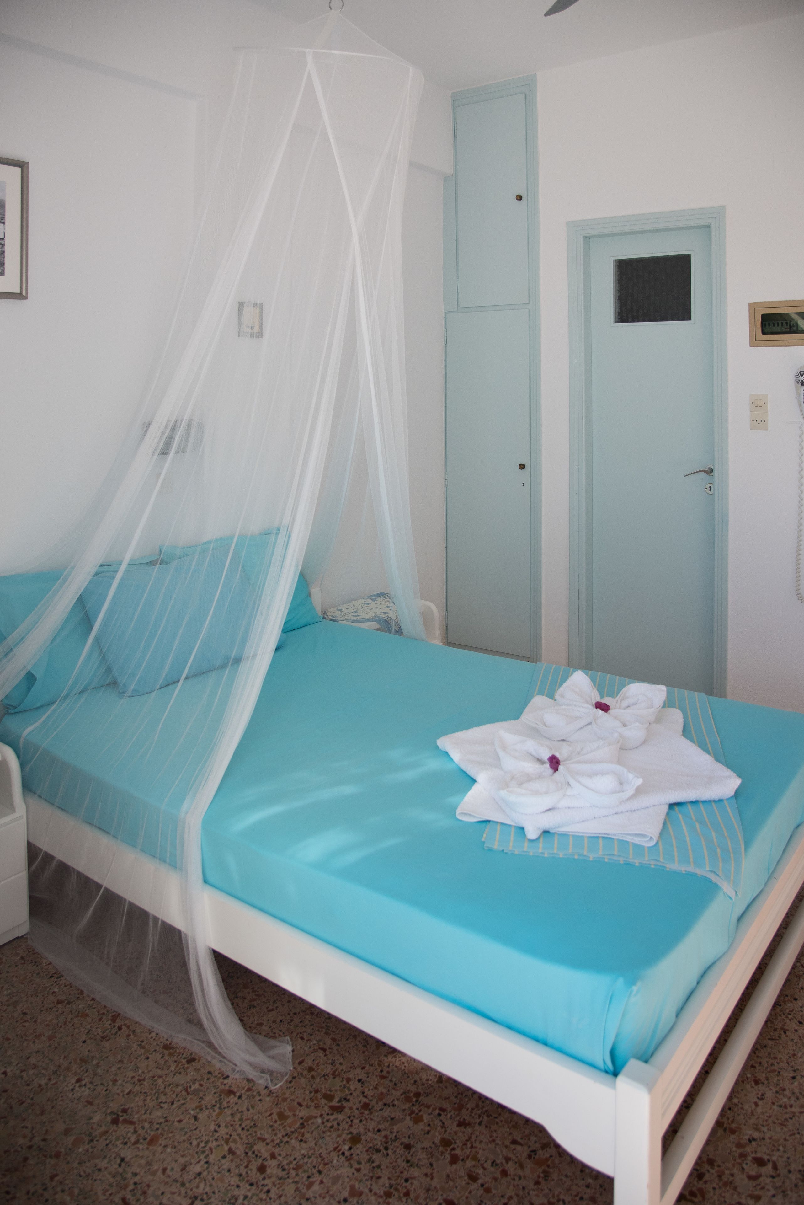 Una cama con colcha azul y almohadas blancas, una mesita de noche blanca, un dosel blanco y una puerta azul claro.