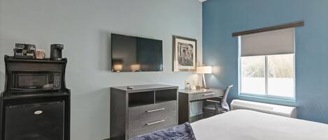 Quarto de hotel com cama, escrivaninha, cadeira, micro-ondas, frigorífico, televisão e janela com persianas.