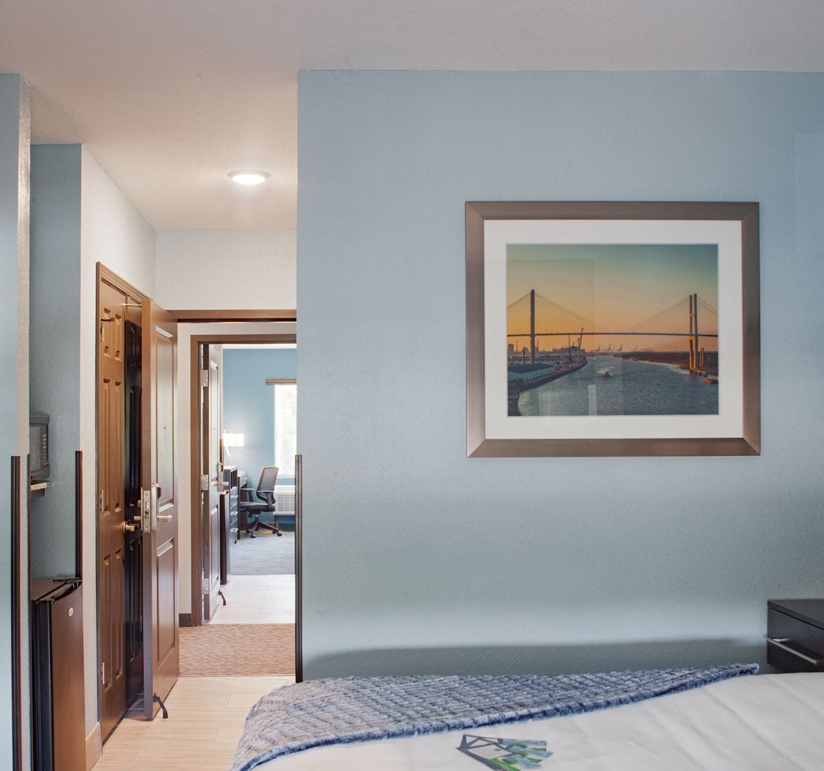 A hotel room with a bed, a framed picture of a bridge, a desk, and a chair.