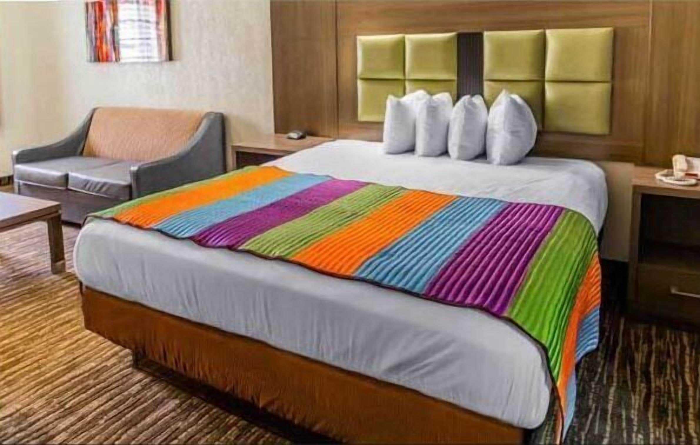 A hotel room with a large bed, a grey armchair, a wooden headboard, and a bedside table with a lamp.