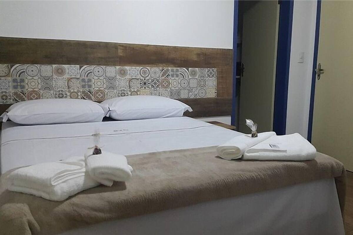 Ein Hotelzimmer mit Bett, Kissen, einem Kopfteil mit Muster sowie Handtüchern und einer Tür mit blauer Linie.