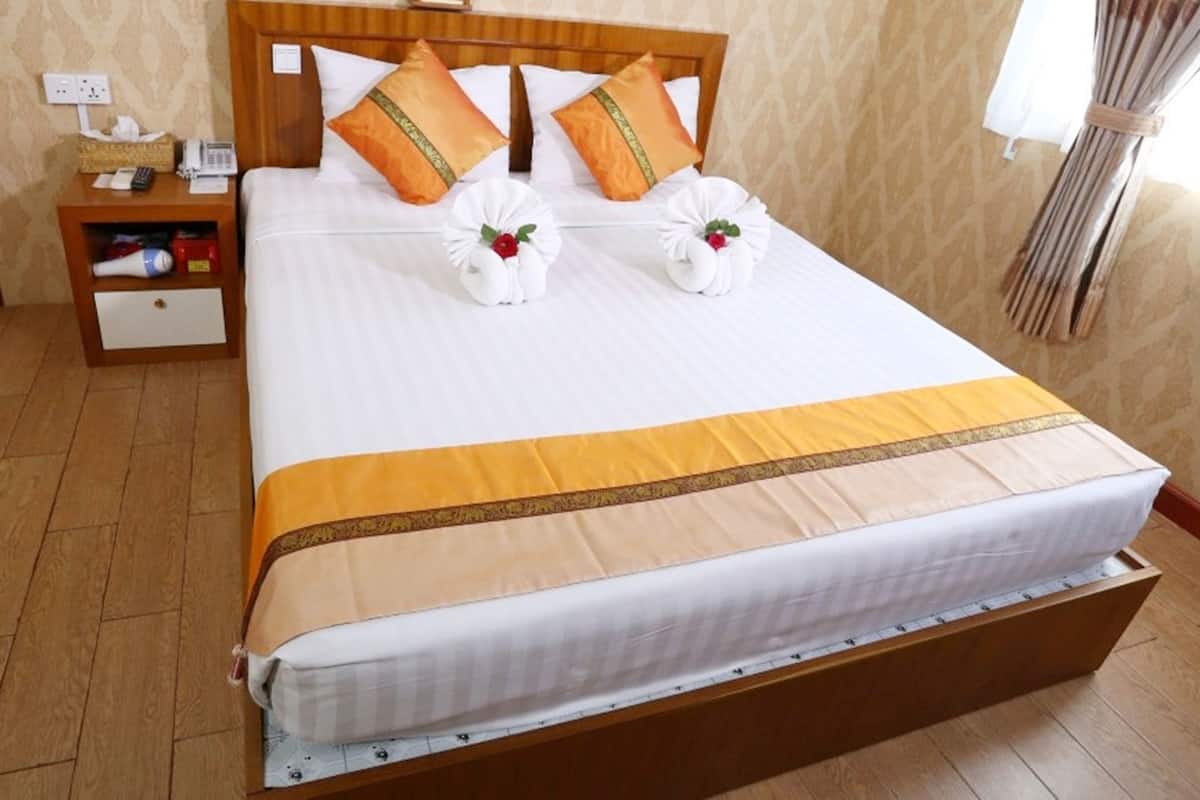 Une chambre d’hôtel avec un grand lit, une tête de lit en bois, deux oreillers, une table de chevet avec une lampe et une petite table de nuit avec un téléphone et une horloge.