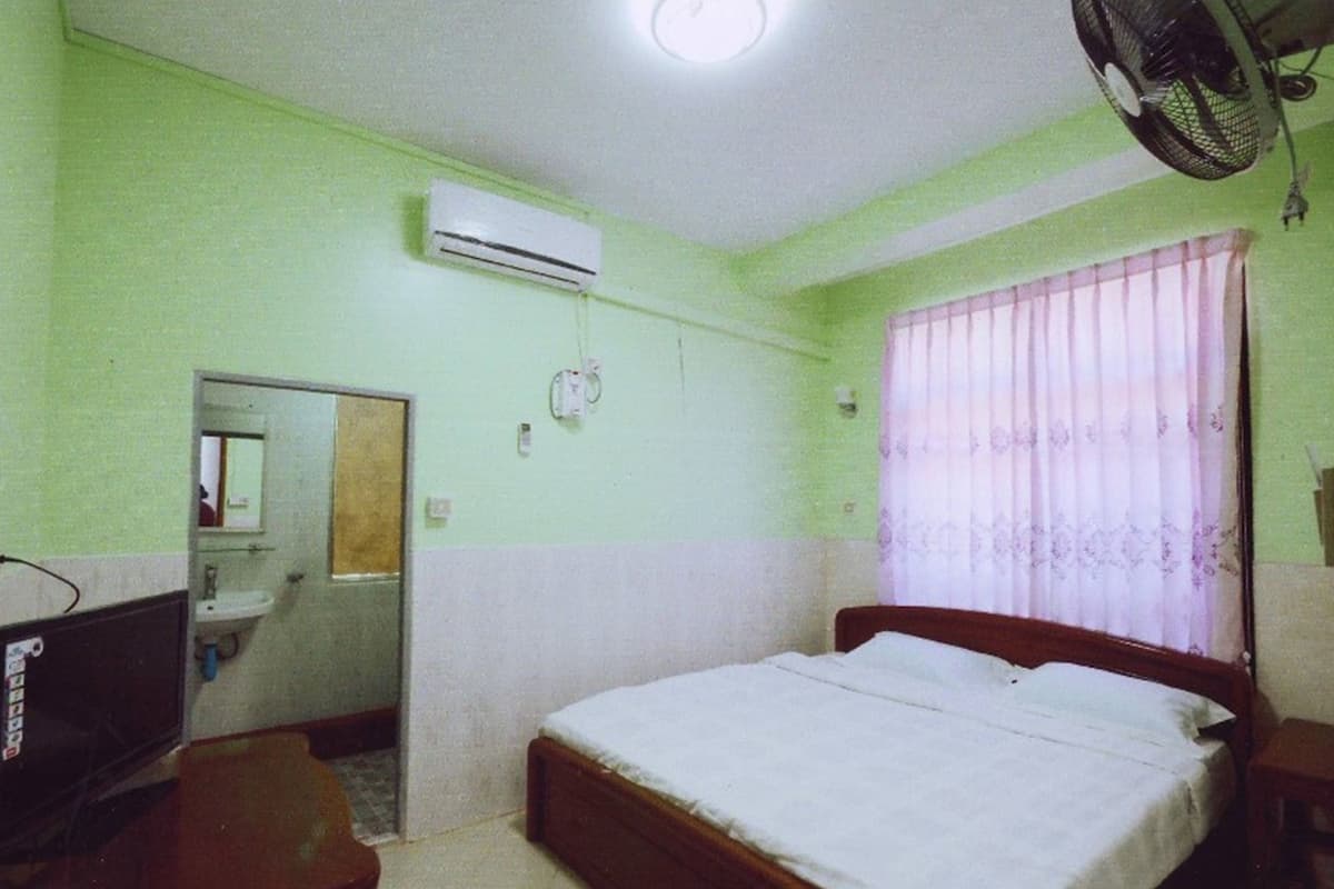 Une chambre à coucher comprenant un lit, une table de chevet, un miroir, un appareil de climatisation, une fenêtre avec des rideaux et un ventilateur.