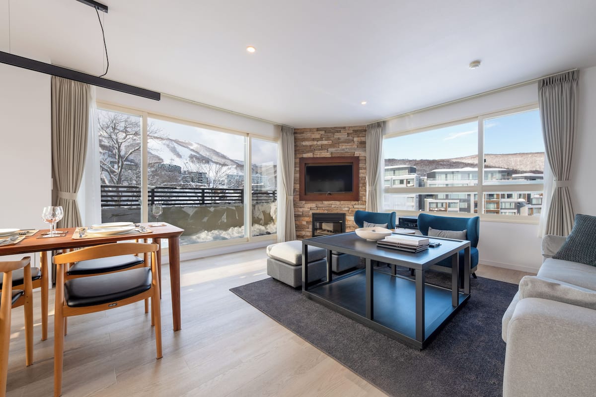 A modern living room with a fireplace, a dining area, and large windows offering a view of a snowy landscape.