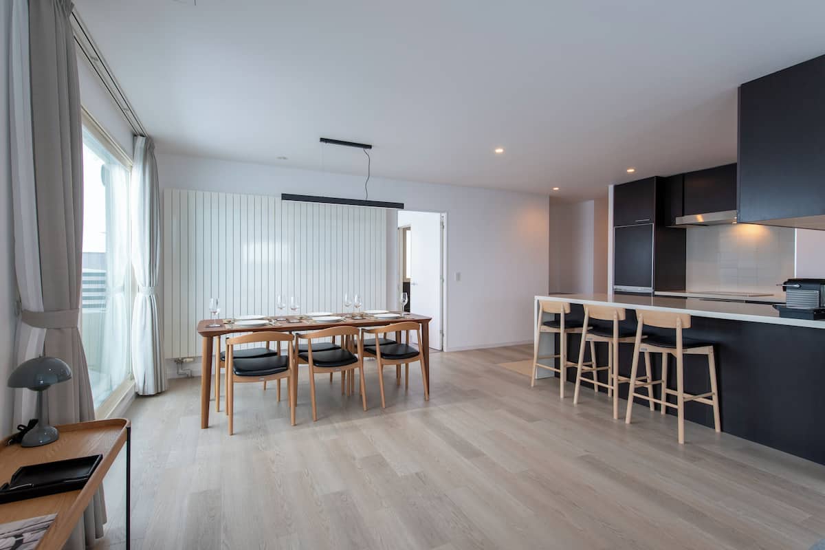 A modern kitchen with a dining area, featuring a wooden table and chairs, a bar area with stools, and a large window with curtains.