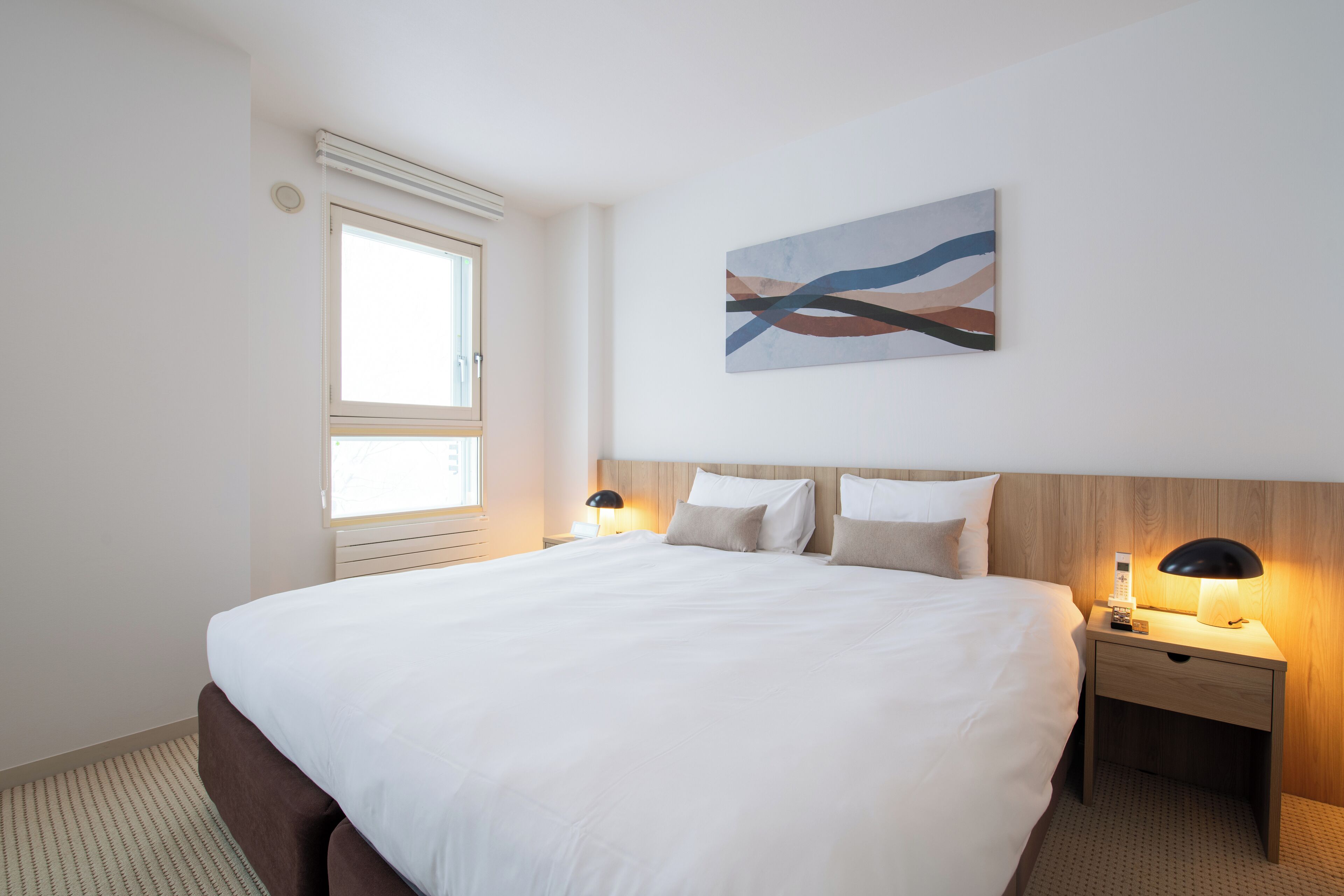 Habitación de hotel con una cama grande, dos mesitas de noche con lámparas, una ventana y un cuadro abstracto colgado en la pared.