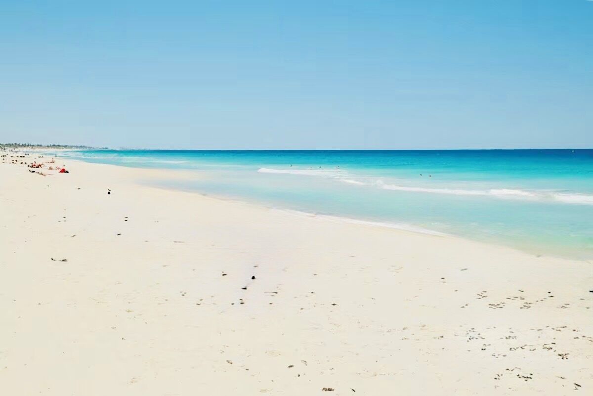 Une vaste plage de sable fin, avec une eau d’un bleu limpide et quelques personnes aperçues au loin.