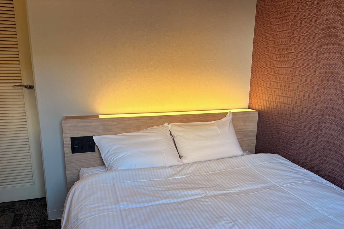 A neatly made bed with white linens and pillows, a headboard with a light fixture, and a plain wall with a textured pattern to the right.
