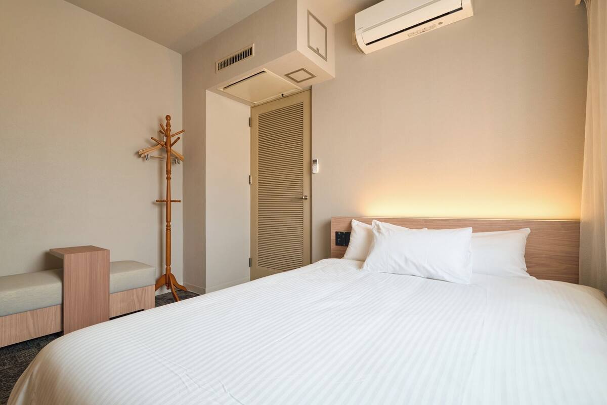 A hotel room with a bed, a bench, a coat stand, an air conditioning unit, and a door with a louvered design.