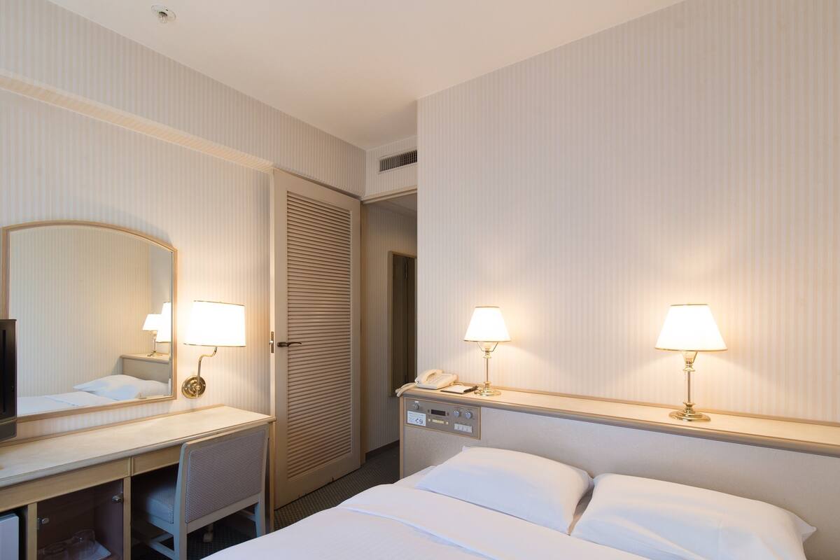 A hotel room with a bed, bedside lamps, a desk with a mirror, a telephone, and a minibar.