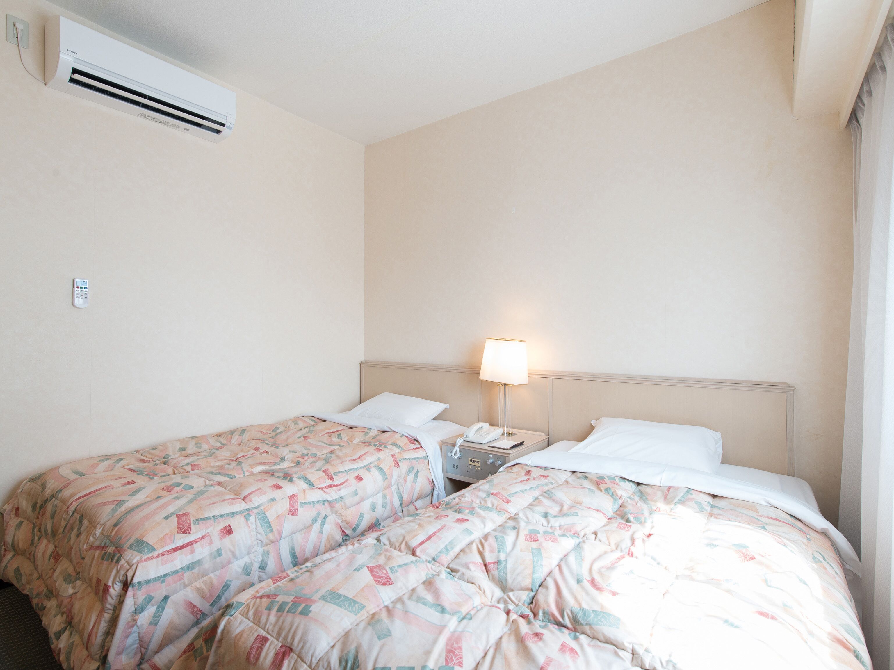 A hotel room with two beds, a bedside table with a lamp, and an air conditioning unit on the wall.