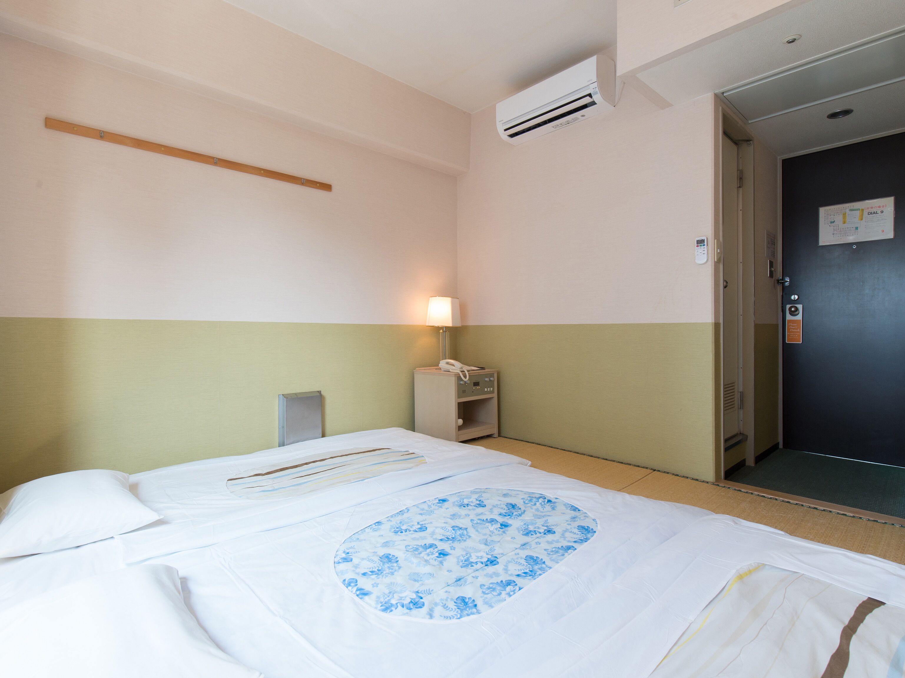 A hotel room with a bed, a bedside table with a lamp, an air conditioning unit, and a door with a sign.