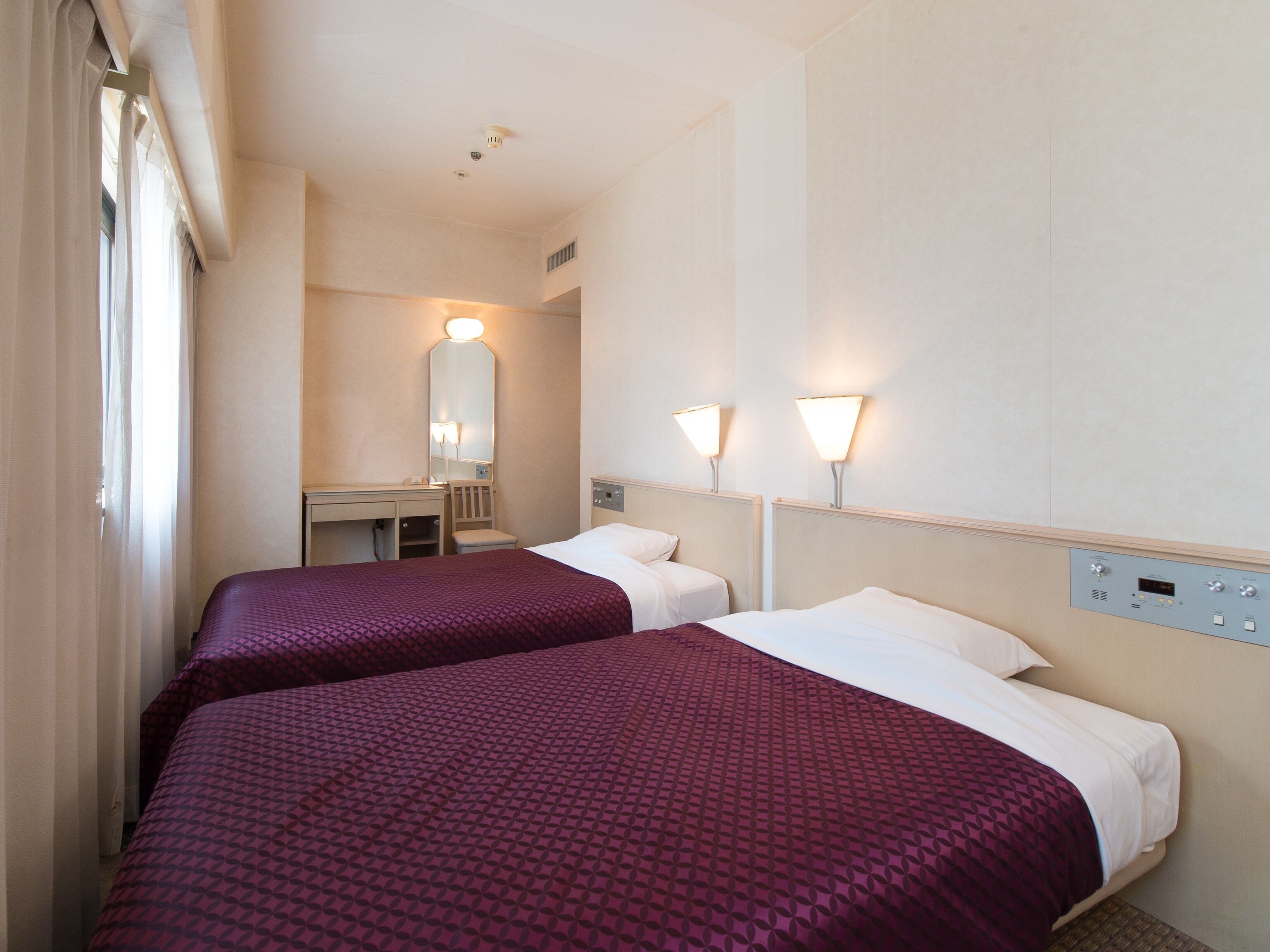 A hotel room with two beds, a desk, a mirror, and wall-mounted lights.