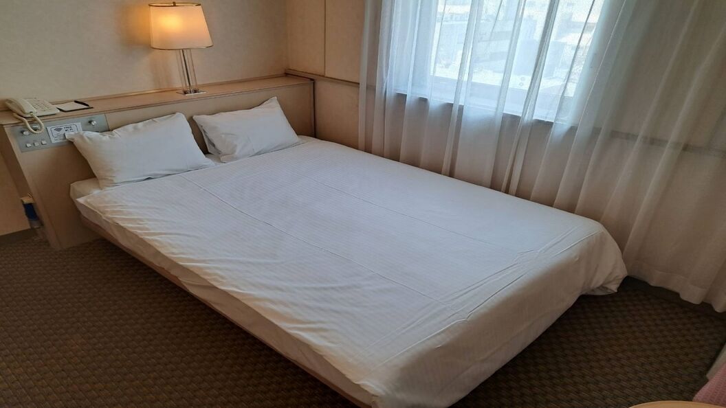 Una camera d'albergo con un letto, un telefono sul comodino, una lampada e una finestra con tende leggere.