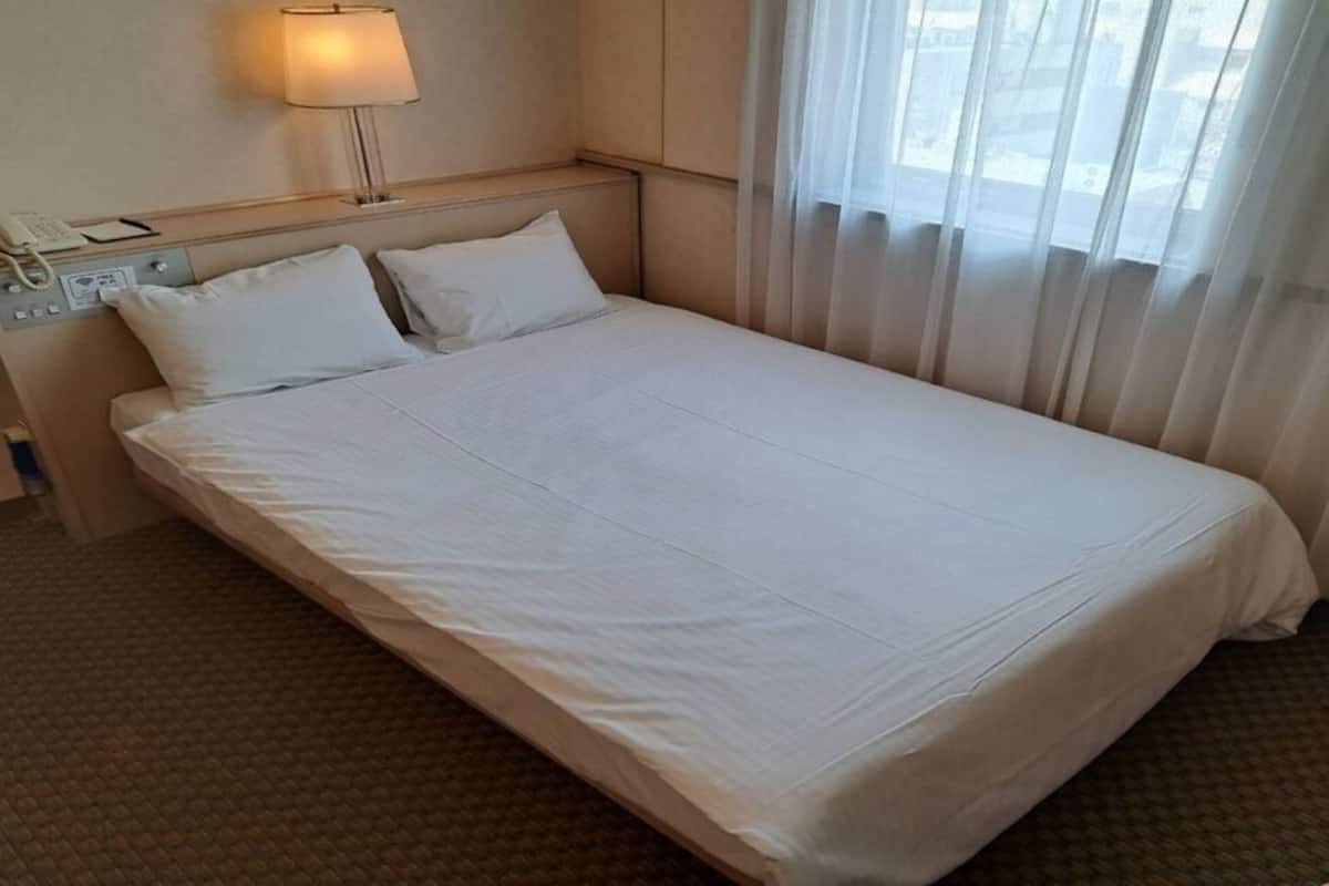 A hotel room with a bed, a bedside telephone, a lamp, and a window with sheer curtains.
