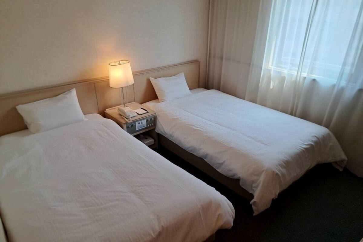 A hotel room with two single beds, a bedside table with a lamp, and a window with sheer curtains.