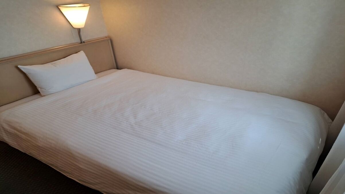 A single bed with a headboard, a wall-mounted lamp, and a plain wall.
