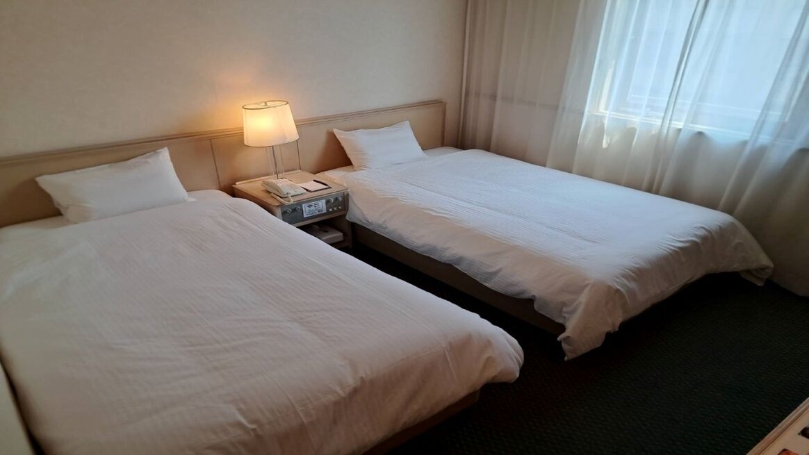 Una camera d'albergo con due letti, un comodino con una lampada e una finestra con tende.