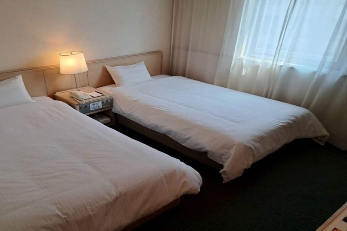 A hotel room with two beds, a bedside table with a lamp, and a window with curtains.