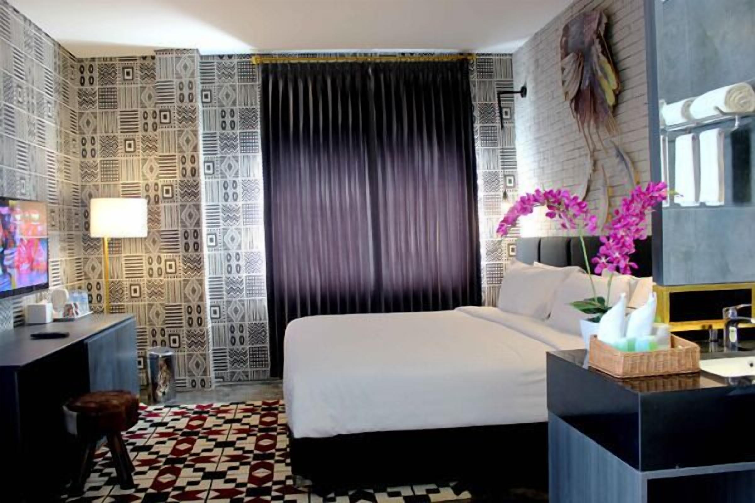 A hotel room with a patterned wallpaper, a large bed, a desk with a lamp, a TV, and a bathroom with a mirror and sink.