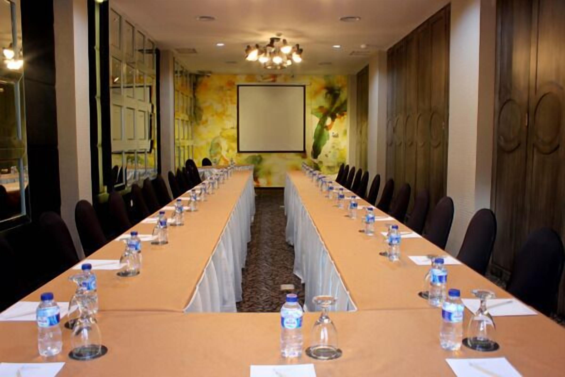 Uma sala de conferência com uma mesa comprida, cadeiras, garrafas de água e uma tela de projetor.