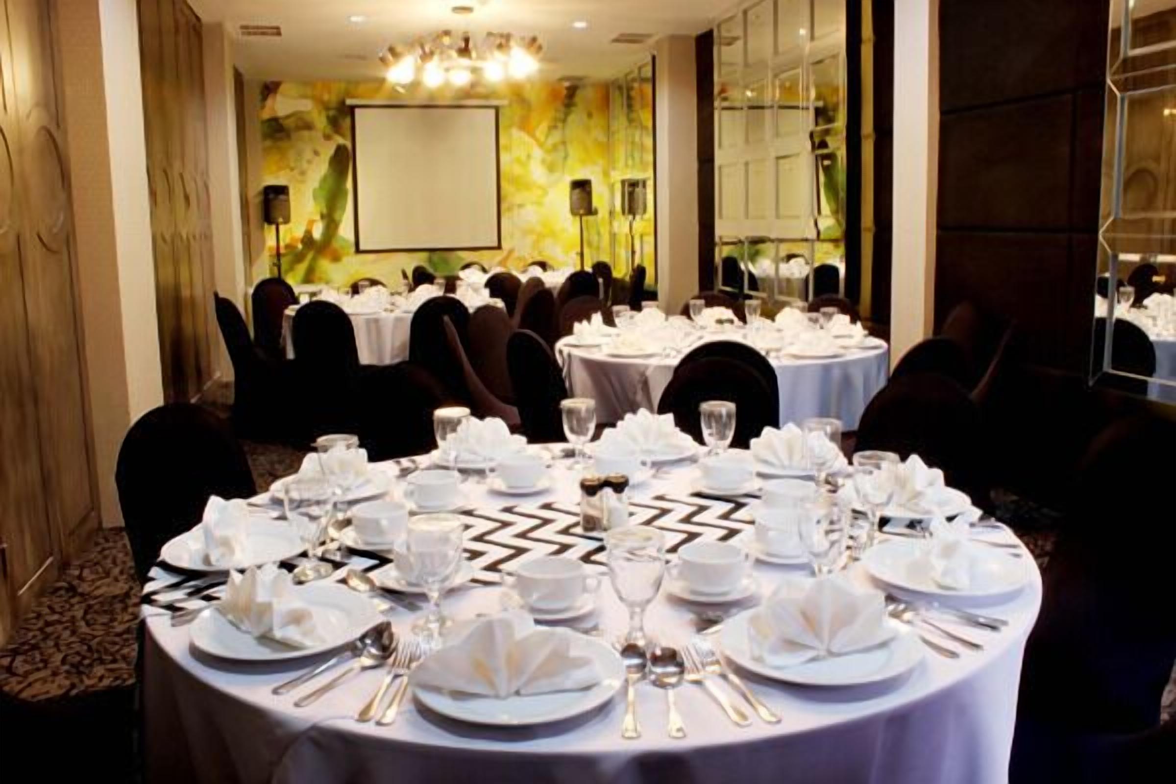Un salón de conferencias con mesas redondas dispuestas para un evento formal, equipado con manteles blancos, platos, cubiertos y vasos.