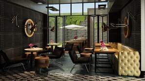 A modern restaurant with a view of greenery through large windows, featuring round tables, leather chairs, and a tufted bench.