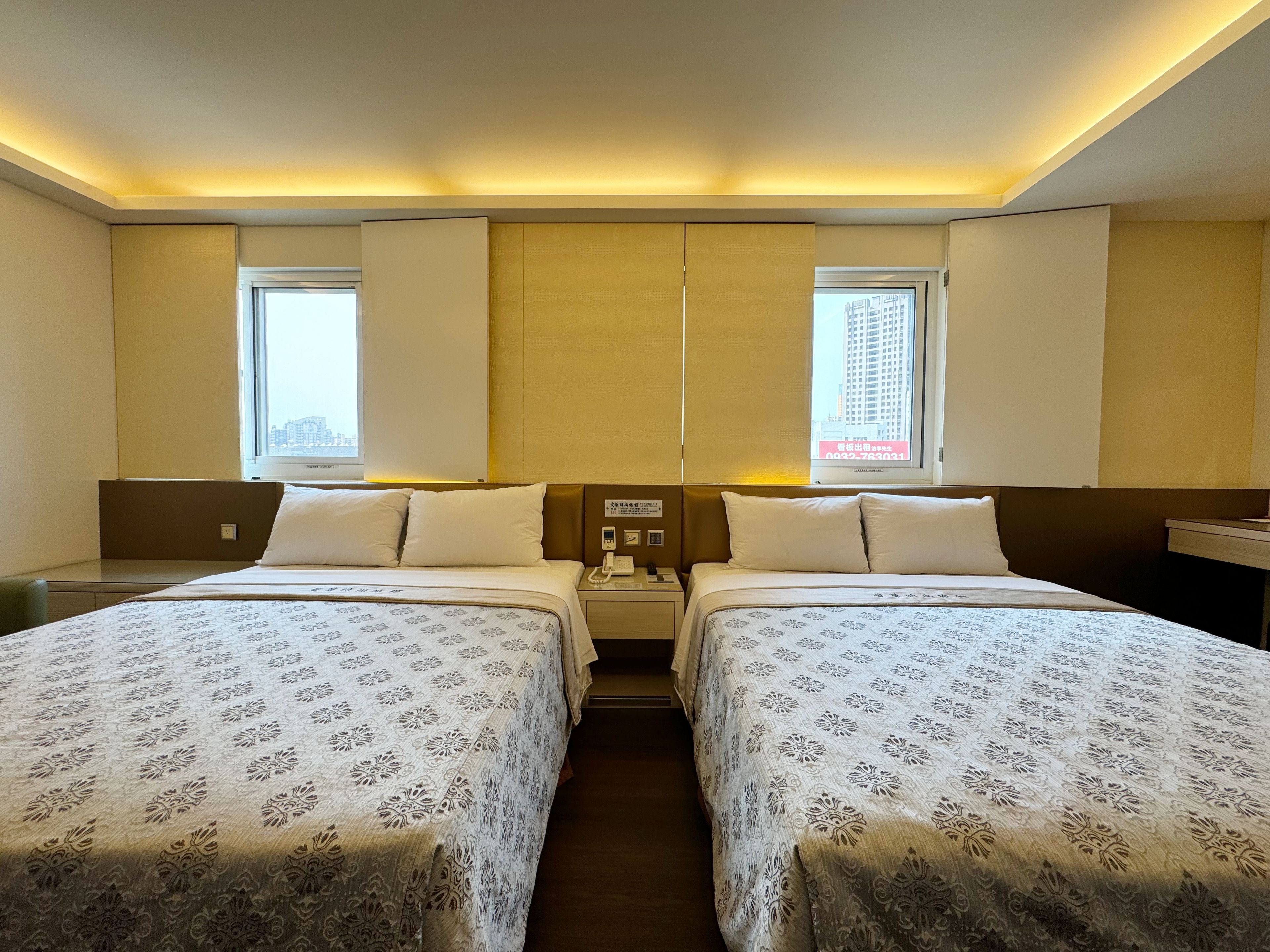 A hotel room with two beds, a bedside table, a nightstand, a window, and a wall-mounted phone.