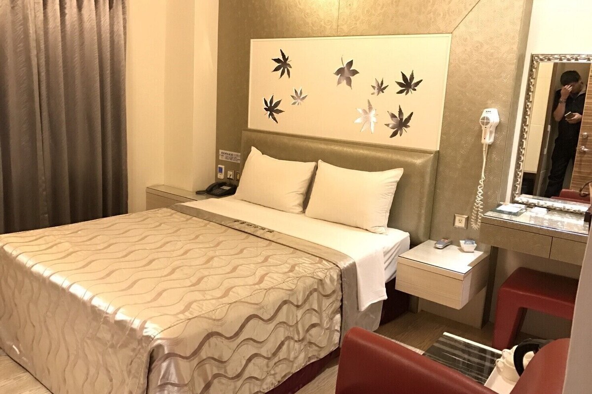 A hotel room with a bed, a bedside table with a phone, a mirror, and a red chair.